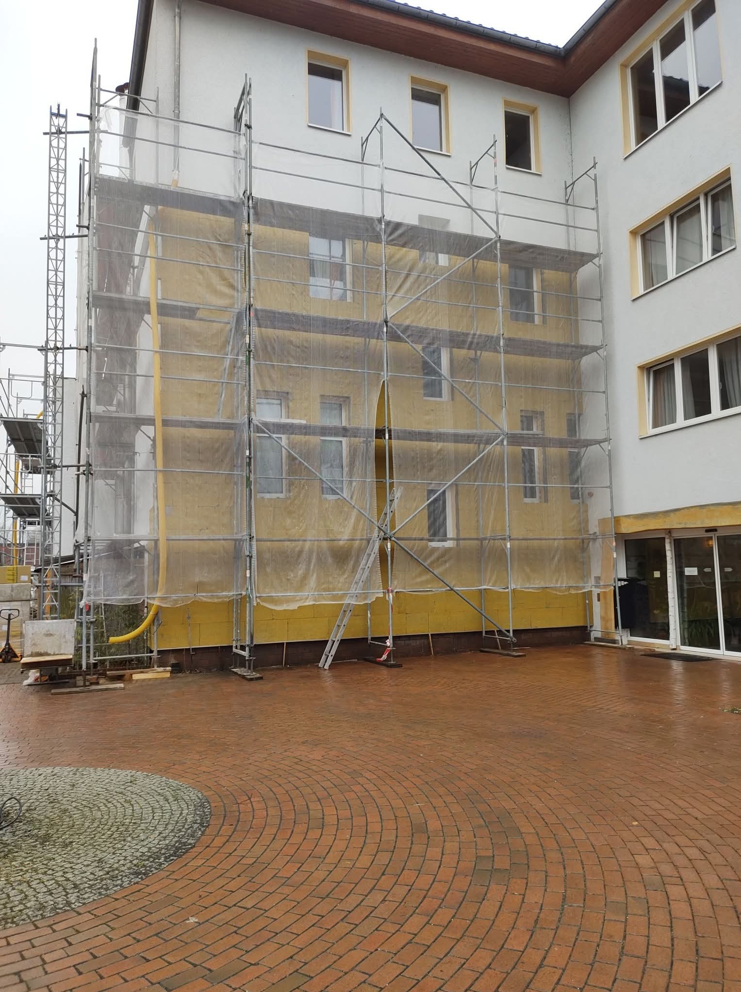 Budynek w trakcie prac elewacyjnych, osłonięty siatką na rusztowaniu. Widoczna żółta elewacja i fragment chodnika z cegły. Prace w toku.