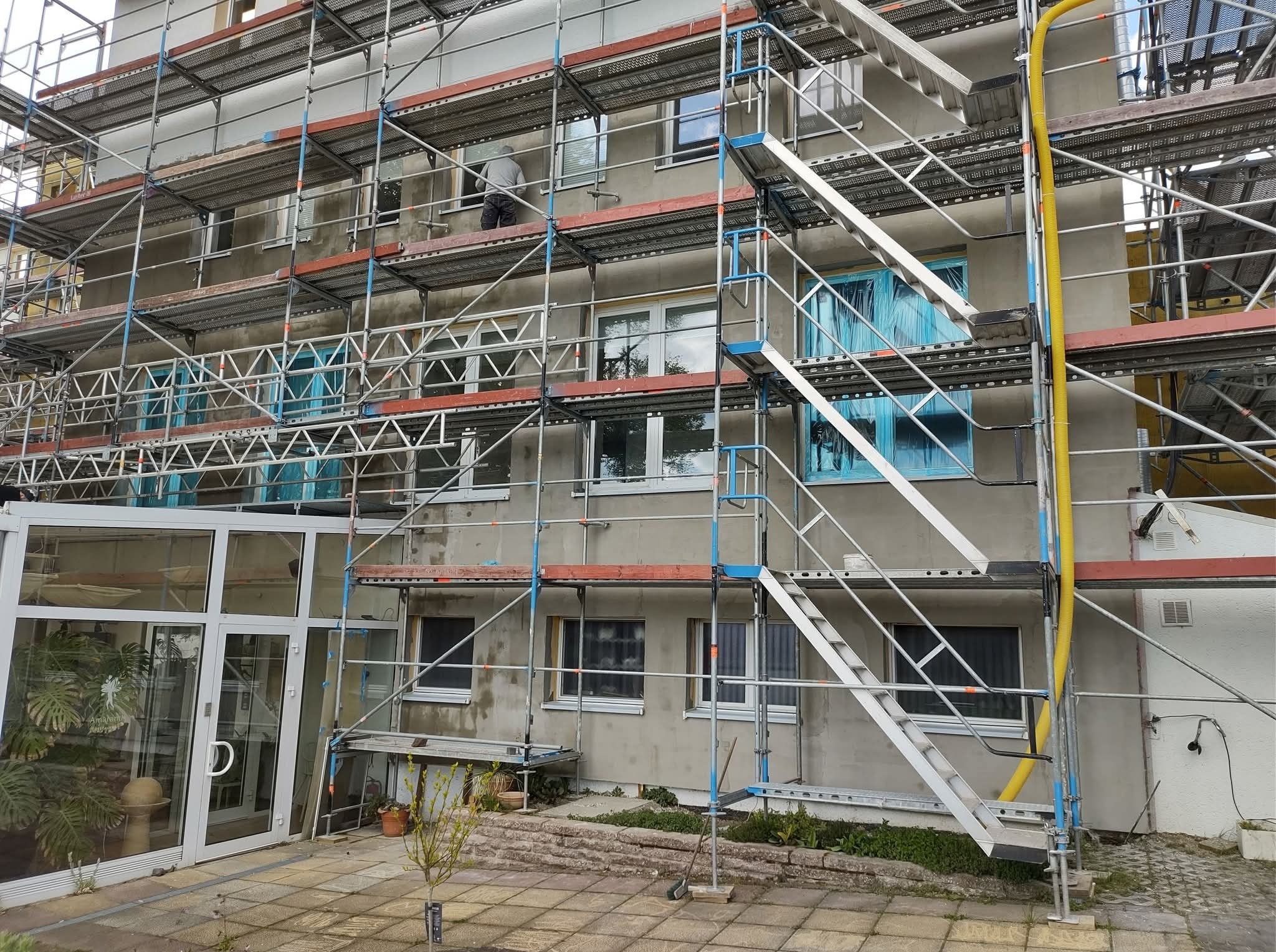 Budynek w trakcie prac elewacyjnych, otoczony rusztowaniami. Widoczny pracownik w trakcie wykonywania prac na wysokości. Okna zabezpieczone folią.