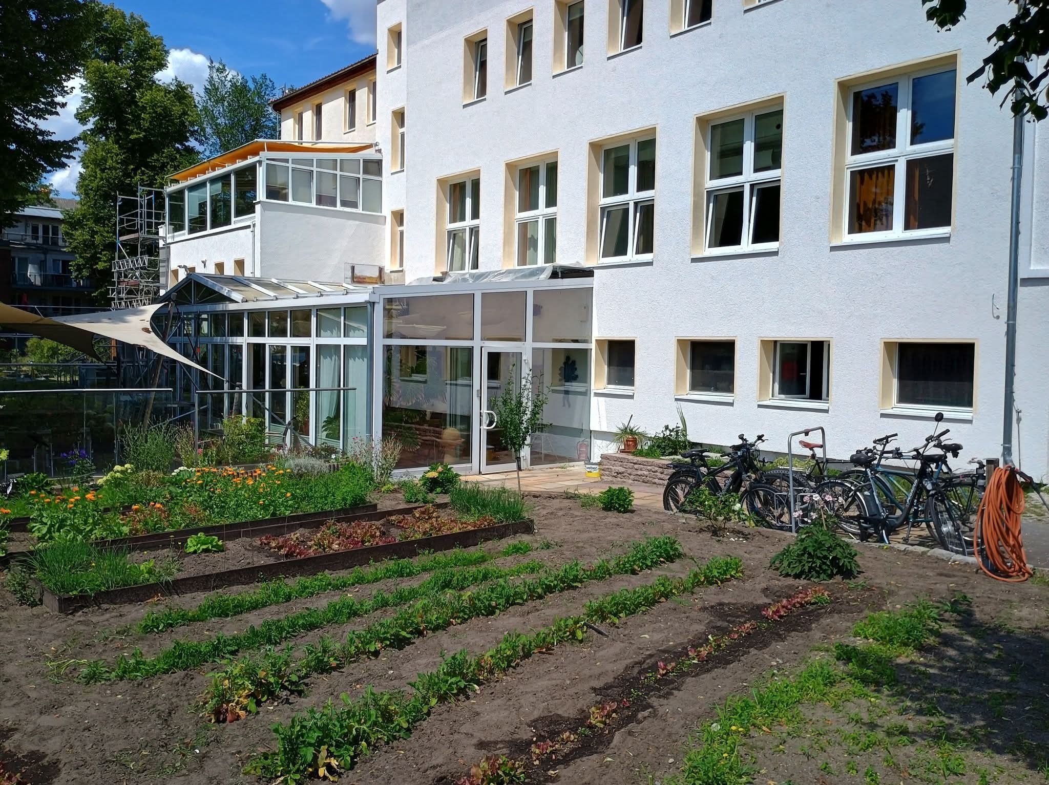 Biały budynek z przeszkloną werandą i ogrodem warzywnym. Widoczne rowy z uprawami, kwiaty, rowery zaparkowane przy ścianie. Słoneczny dzień.