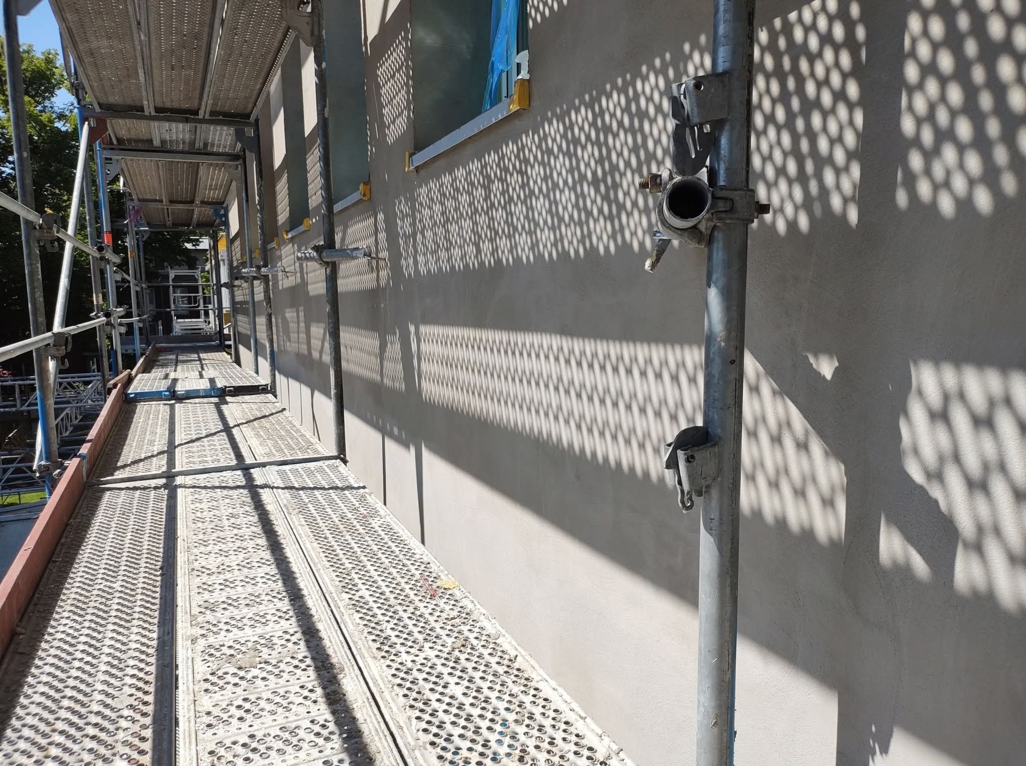 Ściana budynku w trakcie prac wykończeniowych, widoczna z rusztowania. Świeżo nałożona gładź, cień rusztowania na ścianie. Okno zabezpieczone folią.