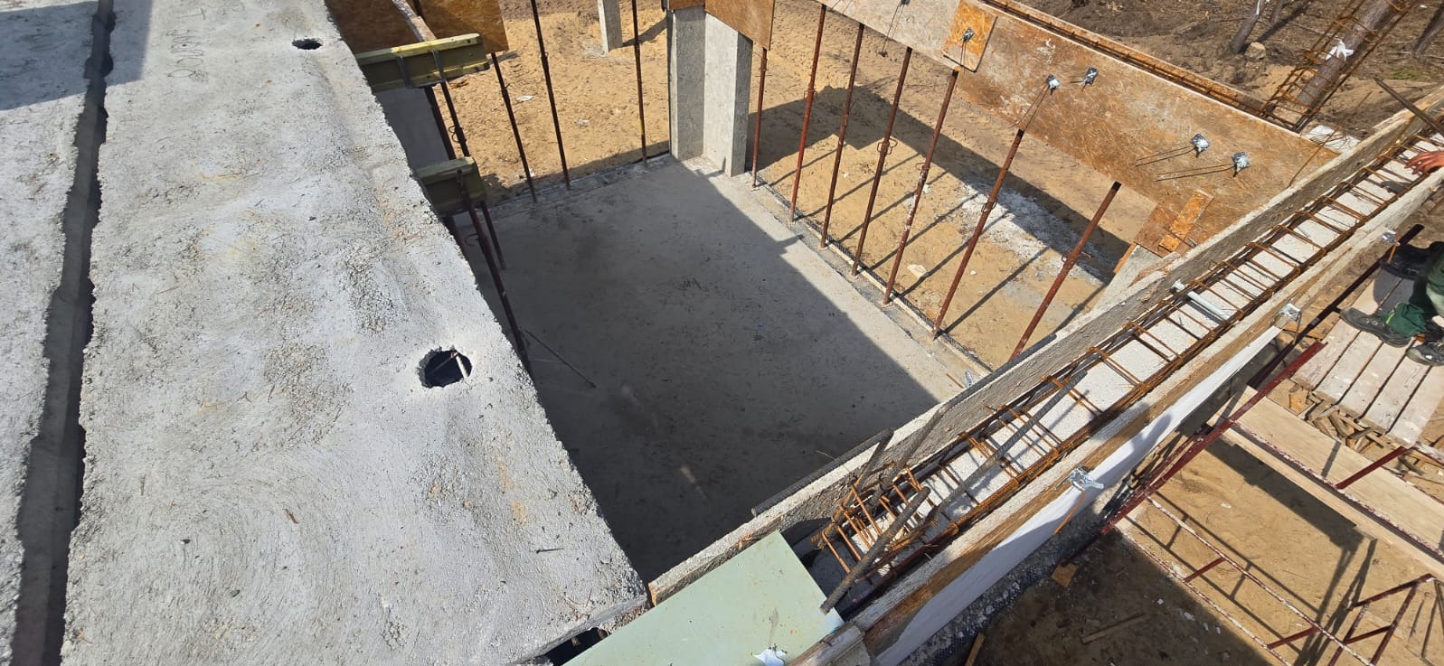 Wykop fundamentów z betonową wylewką, szalunki z desek i zbrojenie. Widok z góry na plac budowy w słoneczny dzień. Elementy konstrukcyjne w surowym stanie.