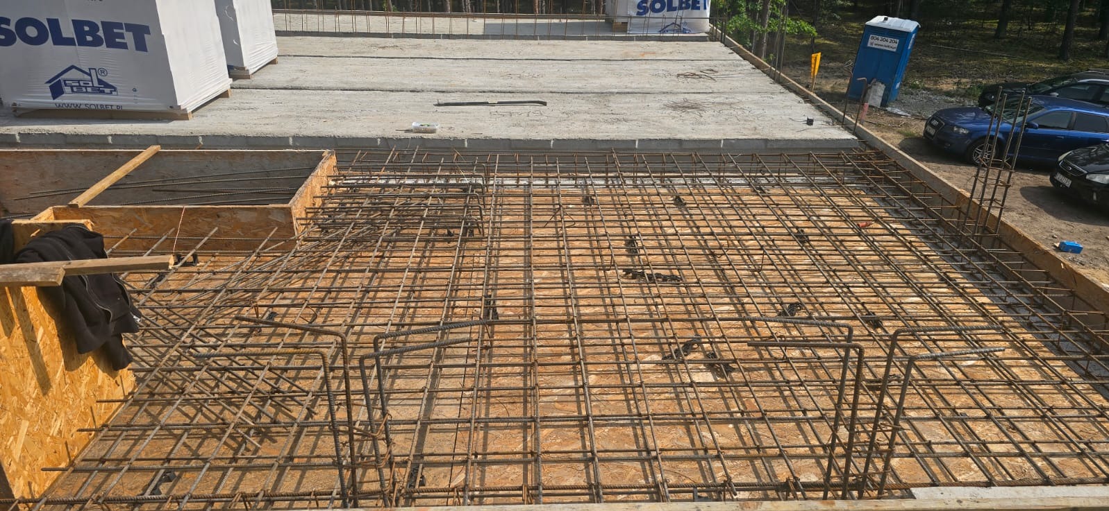 Zbrojenie fundamentów domu jednorodzinnego, widok z góry na siatkę z prętów stalowych w drewnianym szalunku. W tle betonowa płyta i palety z materiałami budowlanymi.