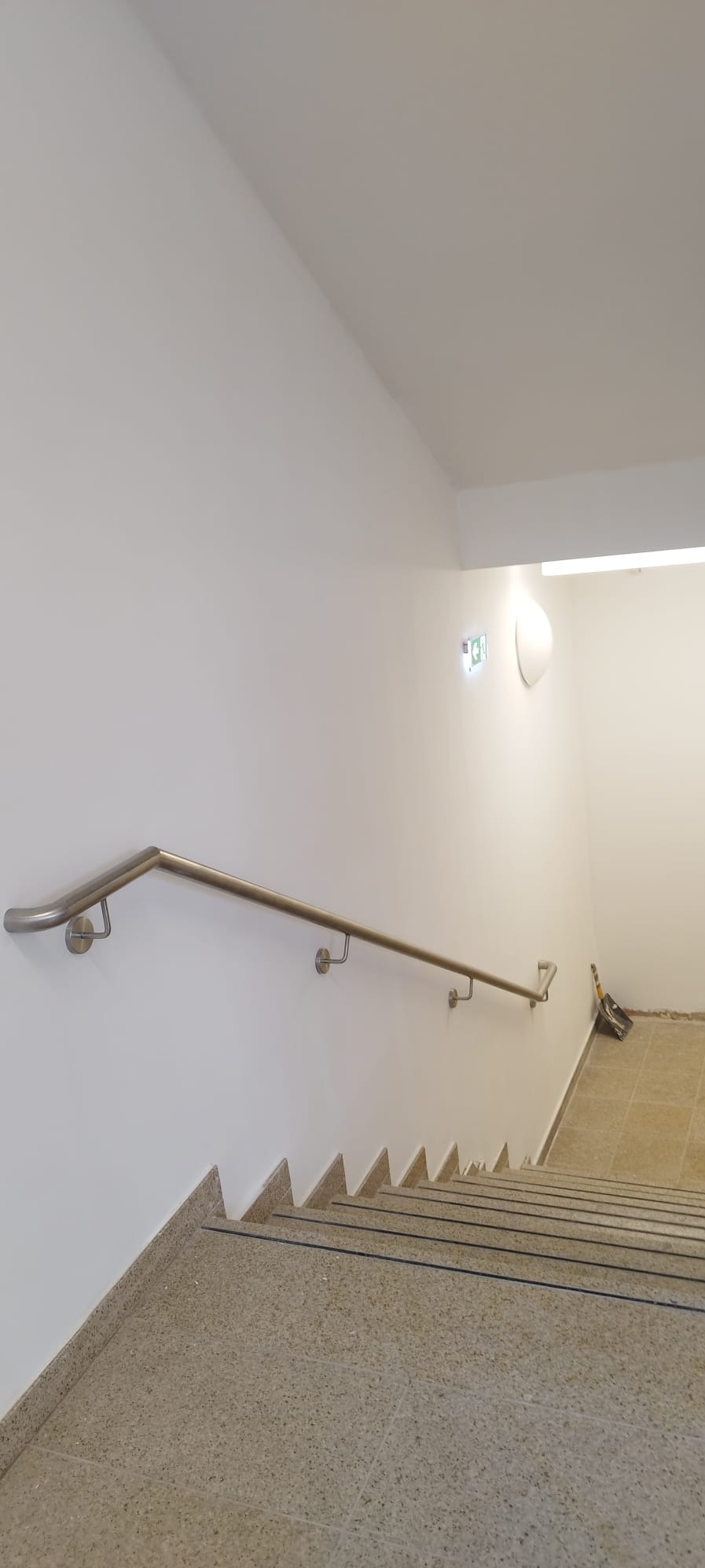 Klatka schodowa po remoncie: jasne ściany, granitowe schody z czarnymi paskami antypoślizgowymi i metalowa poręcz. W tle widoczny sprzęt budowlany.