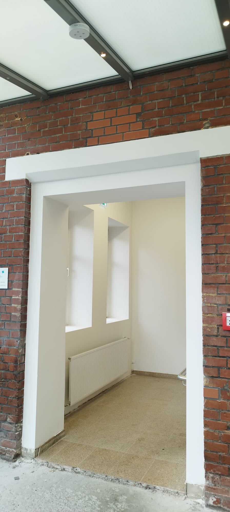 Białe obramowanie drzwi z ceglaną ścianą po bokach. W tle widoczne białe ściany, okna i grzejnik. Na suficie widoczny system oświetlenia i czujnik.