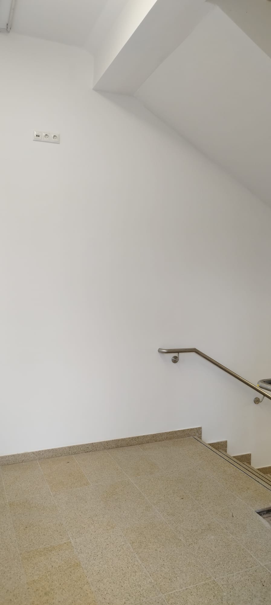Jasne płytki na schodach z metalową poręczą na białej ścianie. Widoczne gniazdka elektryczne. Minimalistyczne wykończenie wnętrza.