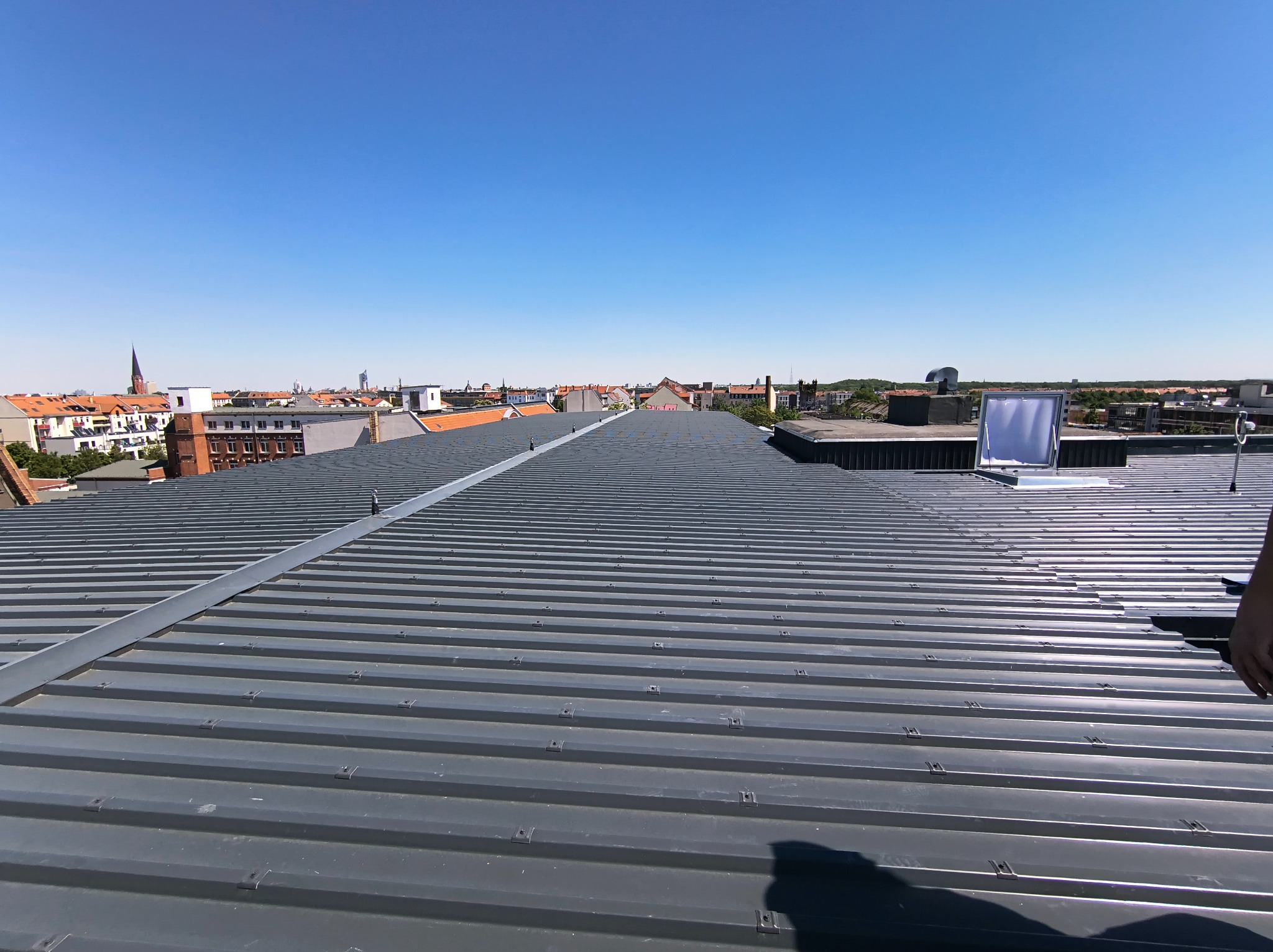 Szary dach z blachy trapezowej, widok z góry na tle zabudowy miejskiej i błękitnego nieba. Widoczne elementy mocujące i fragment ręki w prawym dolnym rogu.