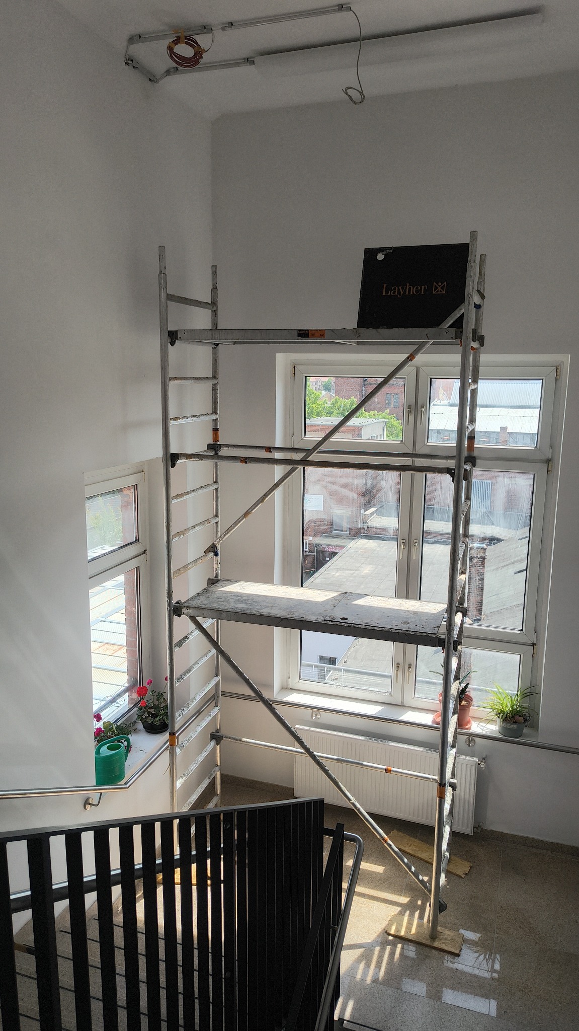 Wnętrze klatki schodowej w trakcie prac. Widoczne rusztowanie Layher, okna z kwiatami i fragment instalacji pod sufitem. Świeżo pomalowane ściany.