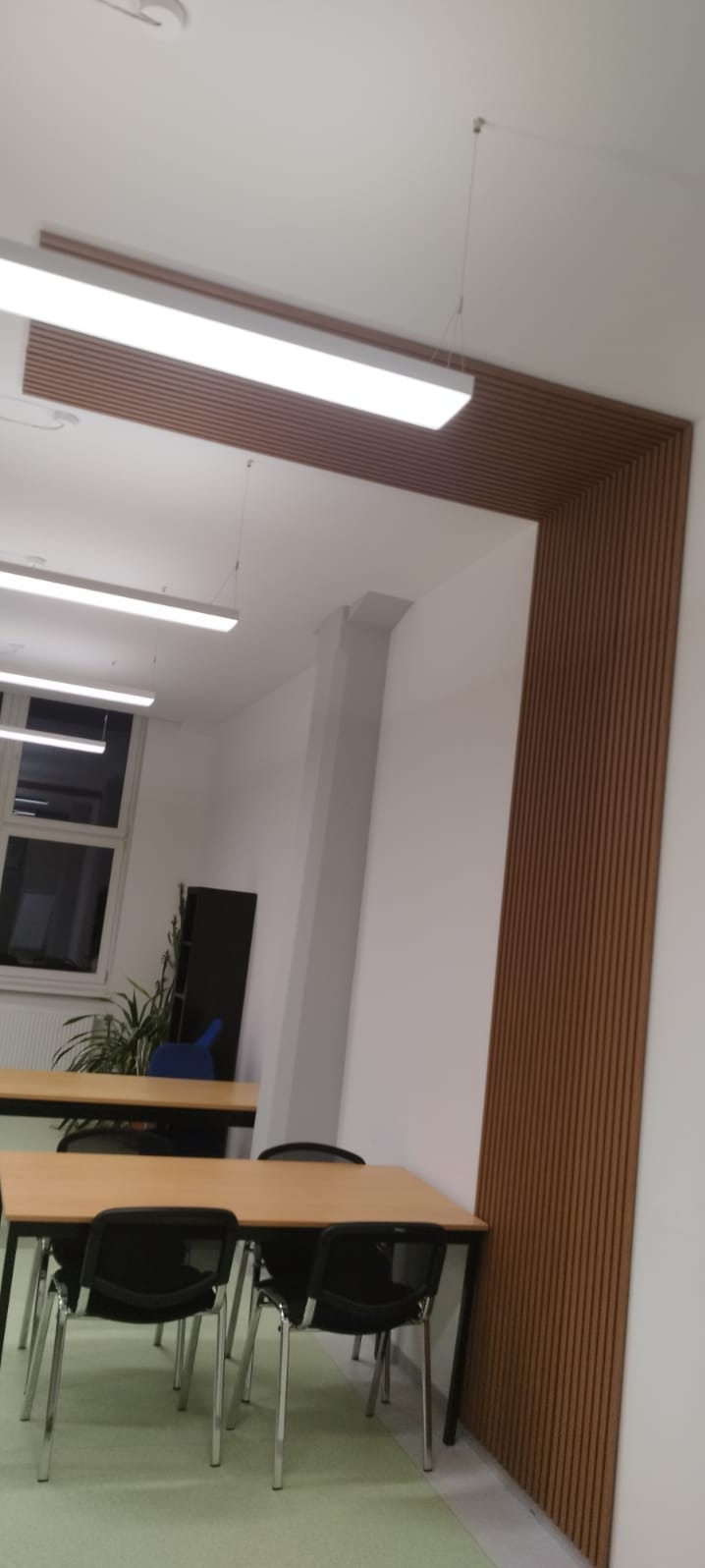 Nowoczesne panele ścienne z drewna w biurze, tworzące dekoracyjny element architektoniczny. Widoczne biurka i krzesła, minimalistyczne oświetlenie.