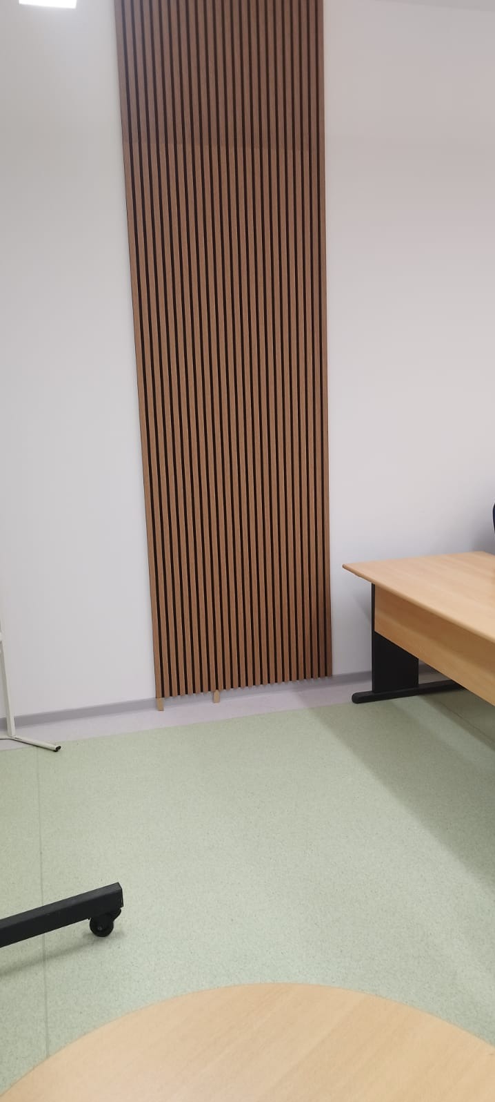 Drewniane lamele dekoracyjne na białej ścianie w biurze, widoczne biurko z blatem w kolorze jasnego drewna i fragment podłogi w odcieniach zieleni.
