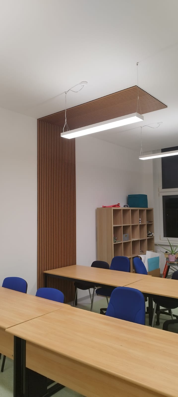 Nowoczesne wykończenie biura: drewniane lamele na ścianie i suficie, podłużne lampy LED, biurka i krzesła w stonowanej kolorystyce. Minimalistyczny design.