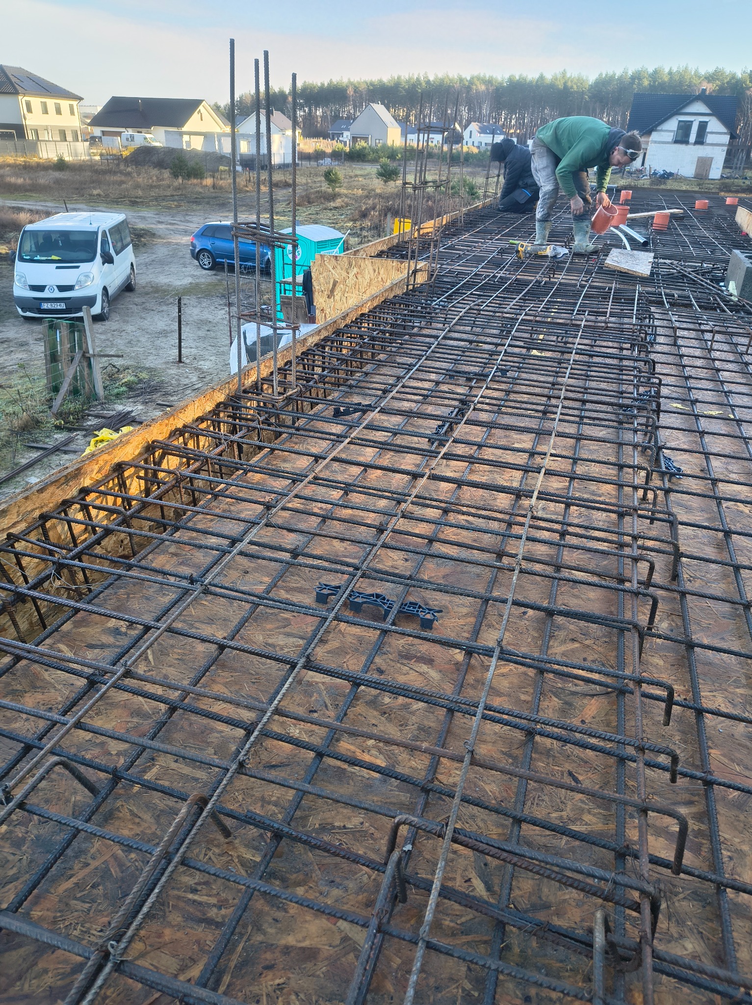 Zbrojenie stropu na deskowaniu z widocznymi pracownikami w trakcie montażu, na tle zabudowy jednorodzinnej i błękitnego nieba. Widoczne narzędzia i elementy instalacji.