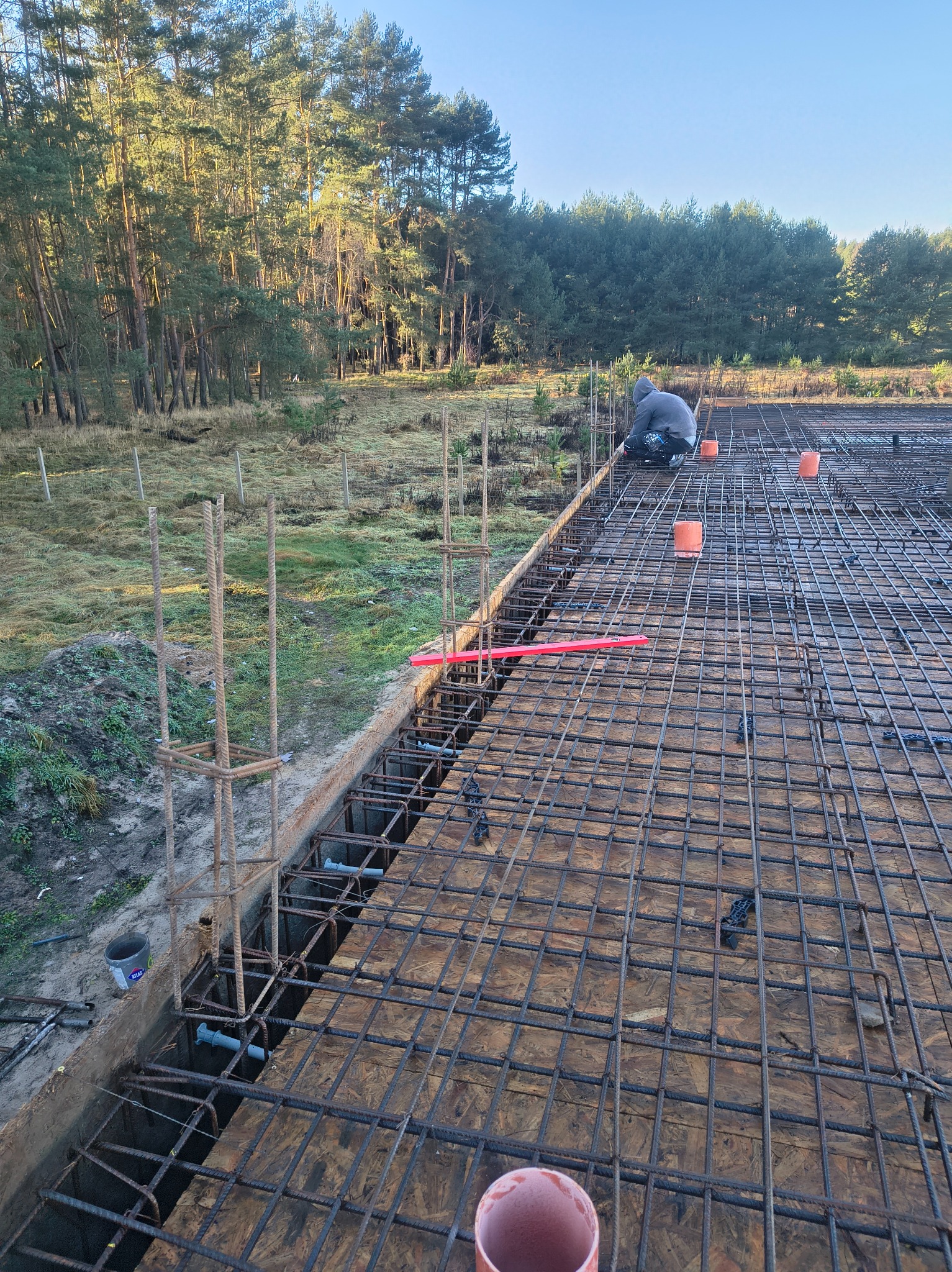 Zbrojenie fundamentów pod budowę domu jednorodzinnego na tle lasu. Widoczne stalowe pręty, szalunek i pracownik w trakcie montażu.