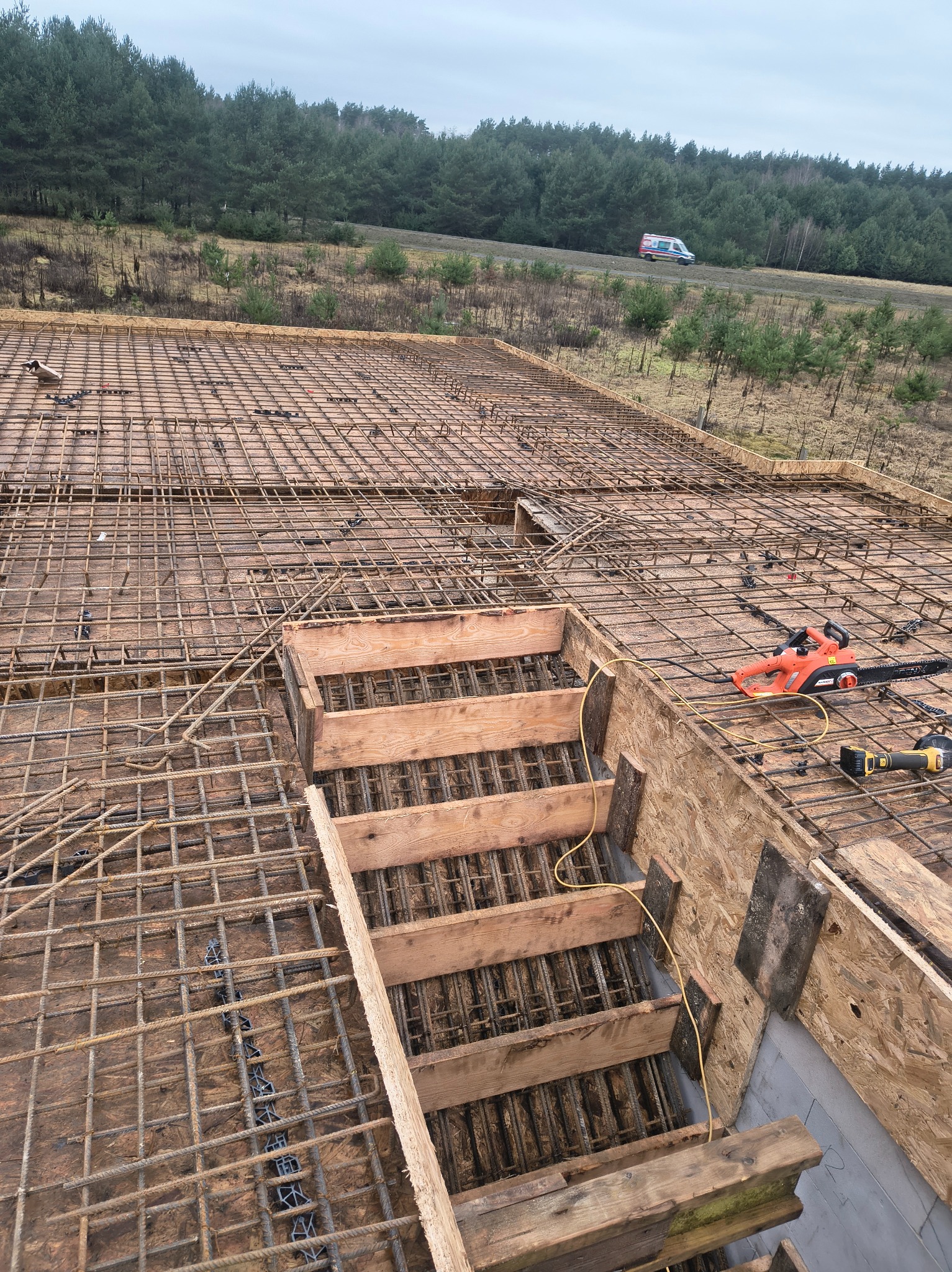 Zbrojenie stropu z drewnianym szalunkiem schodów, widok z góry. Na zbrojeniu leży piła łańcuchowa i szlifierka kątowa. W tle las i przejeżdżający ambulans.