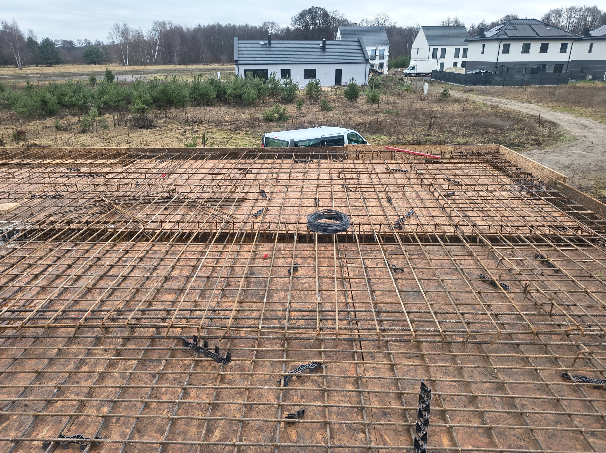 Zbrojenie płyty fundamentowej na budowie domu jednorodzinnego. Widoczne stalowe pręty, dystanse i szalunek. W tle zabudowa mieszkalna i drzewa.
