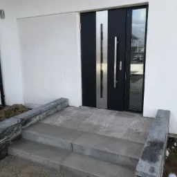 Nowoczesne wejście do domu z czarno-srebrnymi drzwiami i schodami wyłożonymi szarą kostką brukową, obrzeża również z kostki, widoczne ślady prac ziemnych.