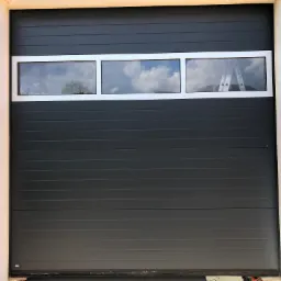 Szara segmentowa brama garażowa z trzema poziomymi oknami, w których odbija się niebo i częściowo drabina, widoczna cegła i surowy beton po bokach.