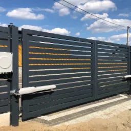 Nowoczesna, pozioma brama przesuwna w kolorze antracytowym z zamontowanymi napędami elektrycznymi, widok z zewnątrz na tle błękitnego nieba z chmurami.