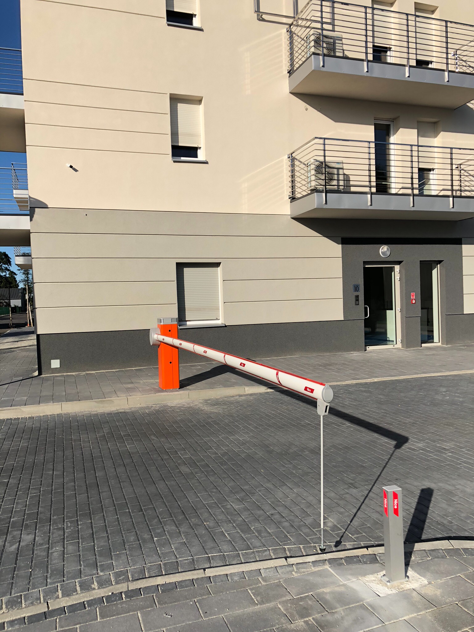 Szlaban automatyczny z pomarańczową obudową, zaporą w kolorze białym z czerwonymi paskami, przed nowoczesnym budynkiem wielorodzinnym z balkonami.