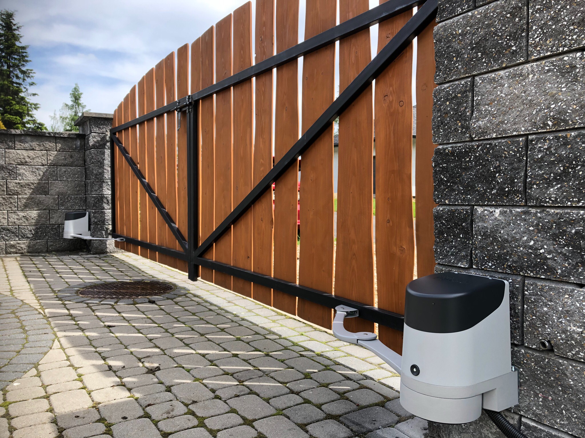 Drewniana brama wjazdowa z czarną metalową ramą i szarym, nowoczesnym napędem zamontowanym na kamiennym słupku z kostki brukowej.