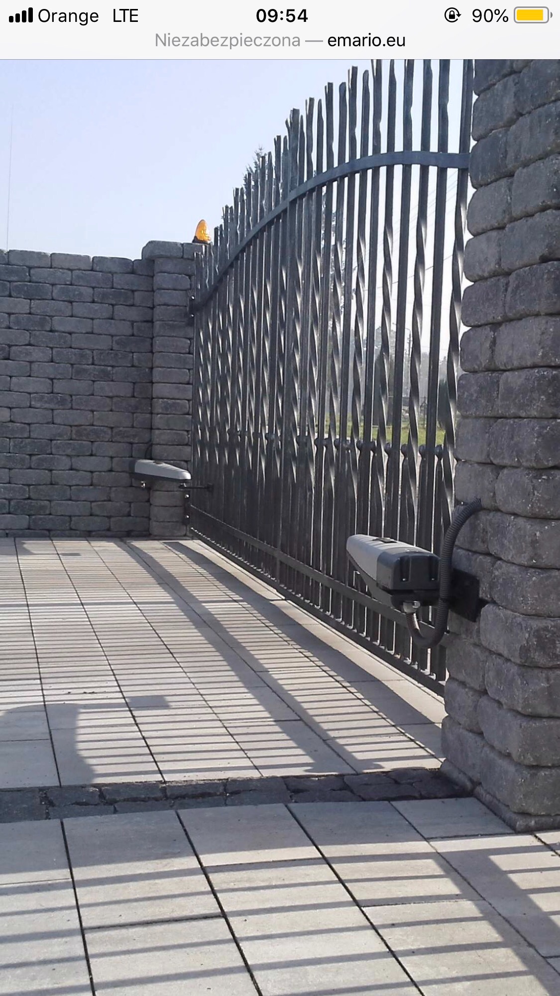 Automatyczna brama wjazdowa z czarnymi, kutymi prętami, zamontowana na słupkach z szarej cegły, z widocznym napędem elektrycznym i cieniem rzucanym przez bramę na szarą kostkę brukową.