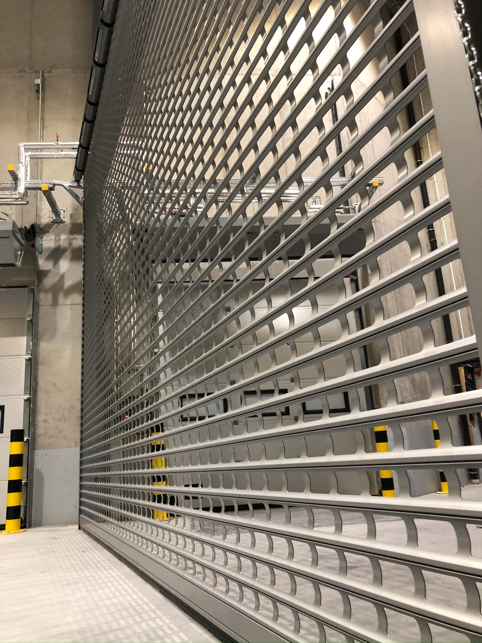 Szara, metalowa brama rolowana z perforowanymi profilami, częściowo otwarta, widoczna instalacja pod sufitem i słupki ostrzegawcze w tle.
