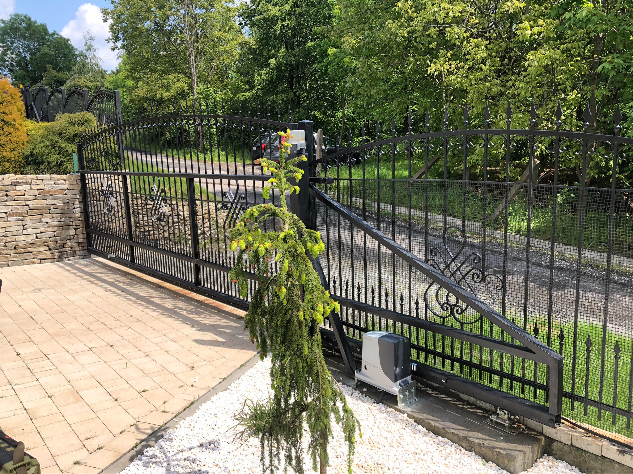 Czarna, metalowa brama wjazdowa z dekoracyjnymi elementami, napęd bramy zamontowany na fundamencie z białych kamieni, w tle droga i zieleń.