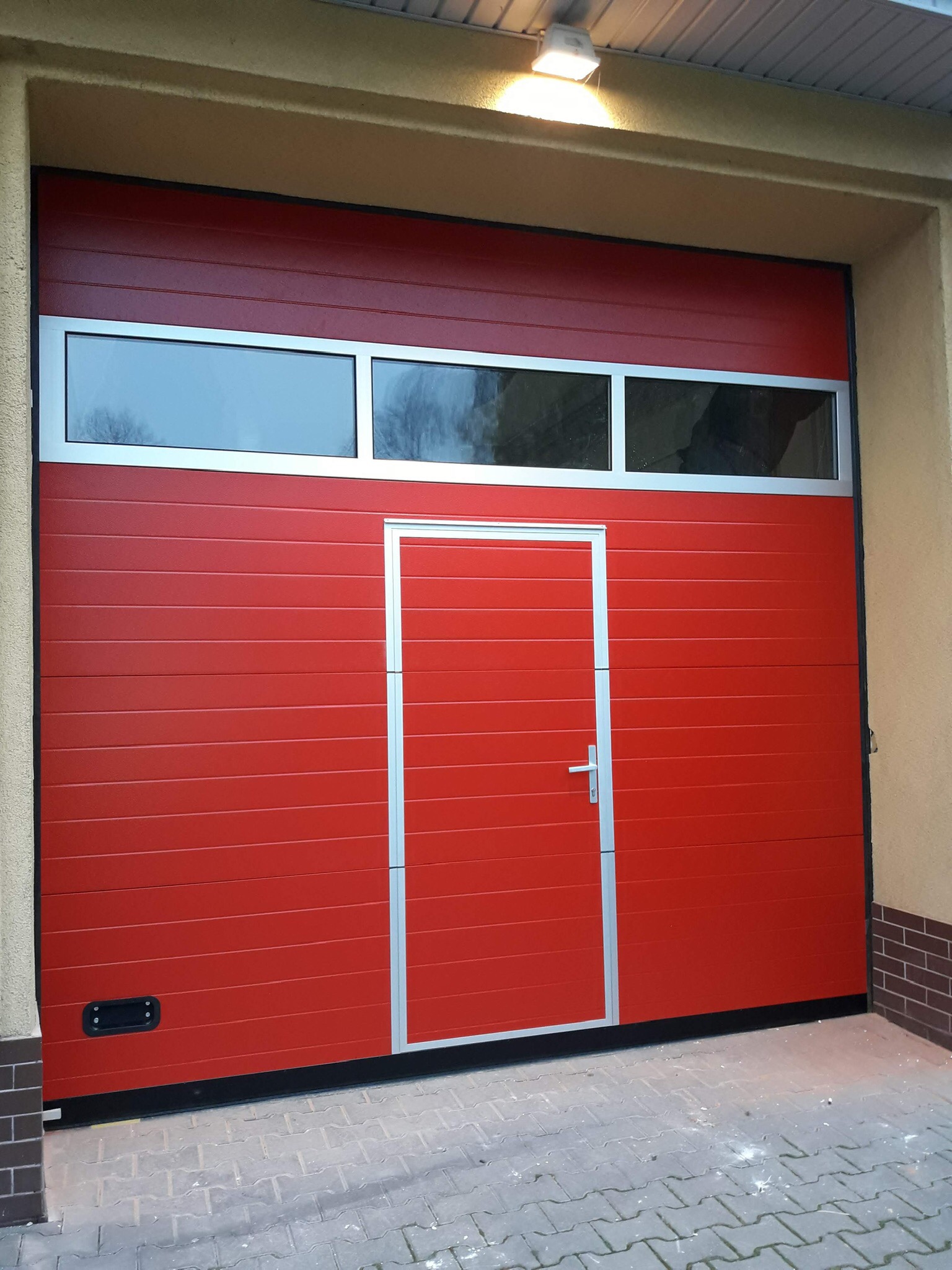 Czerwona segmentowa brama garażowa z trzema oknami i drzwiami wejściowymi, srebrne obramowanie, szara kostka brukowa przed bramą, oświetlenie górne.