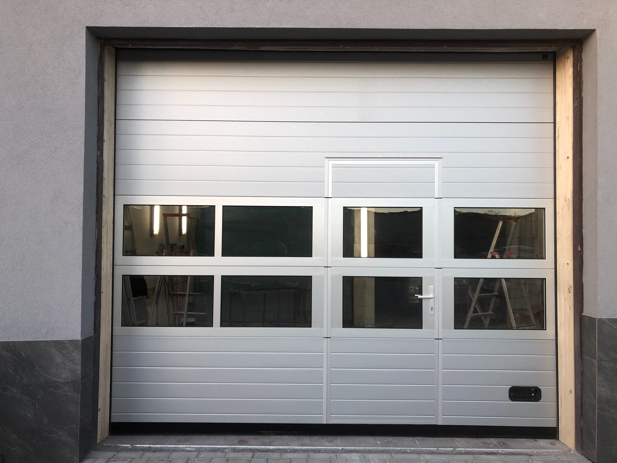 Biała segmentowa brama garażowa z oknami, widoczne drabiny wewnątrz garażu.