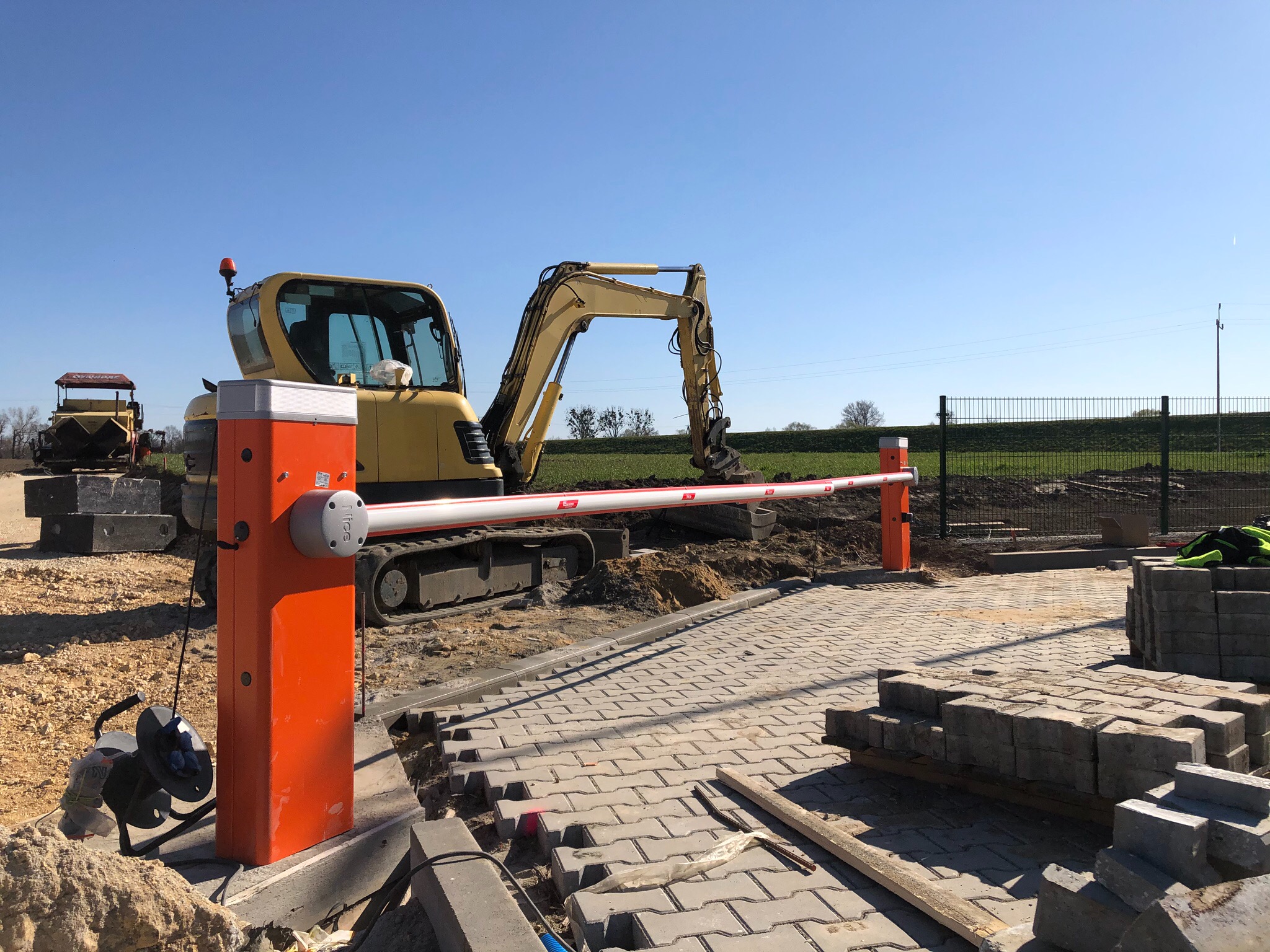 Pomarańczowy szlaban automatyczny z żółtą koparką w tle, kostka brukowa w trakcie układania, plac budowy w słoneczny dzień.