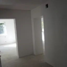 Surowe wnętrze w trakcie prac wykończeniowych; widoczne ściany po nałożeniu gładzi, przygotowane do malowania, z widocznymi otworami okiennymi i drzwiowymi.