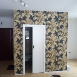 NEW-BUD Norbert Janik - Ściana z tapetą w motywy tropikalne z widocznymi drzwiami przesuwnymi i fragmentem łazienki w tle, pomieszczenie w trakcie prac wykończeniowych, widoczna miotła i narzędzia na podłodze.