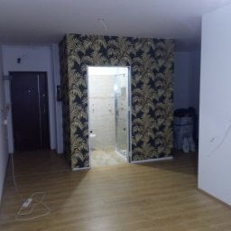 NEW-BUD Norbert Janik - Pokój po remoncie z nową tapetą w palmy na ścianie z wejściem do łazienki, widoczna podłoga laminowana i drzwi wejściowe.