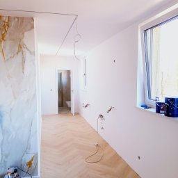 NEW-BUD Norbert Janik - Wnętrze w trakcie wykańczania: parkiet w jodełkę, ściana z marmurowym wzorem, białe ściany z wyprowadzonymi instalacjami, puszki farb na parapecie. Widok na łazienkę.