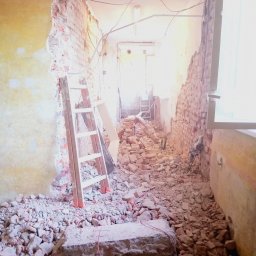 NEW-BUD Norbert Janik - Demontaż ściany z cegły w trakcie remontu. Widoczne gruz, drabina oparta o ścianę i narzędzia. Prace remontowe wewnątrz budynku.