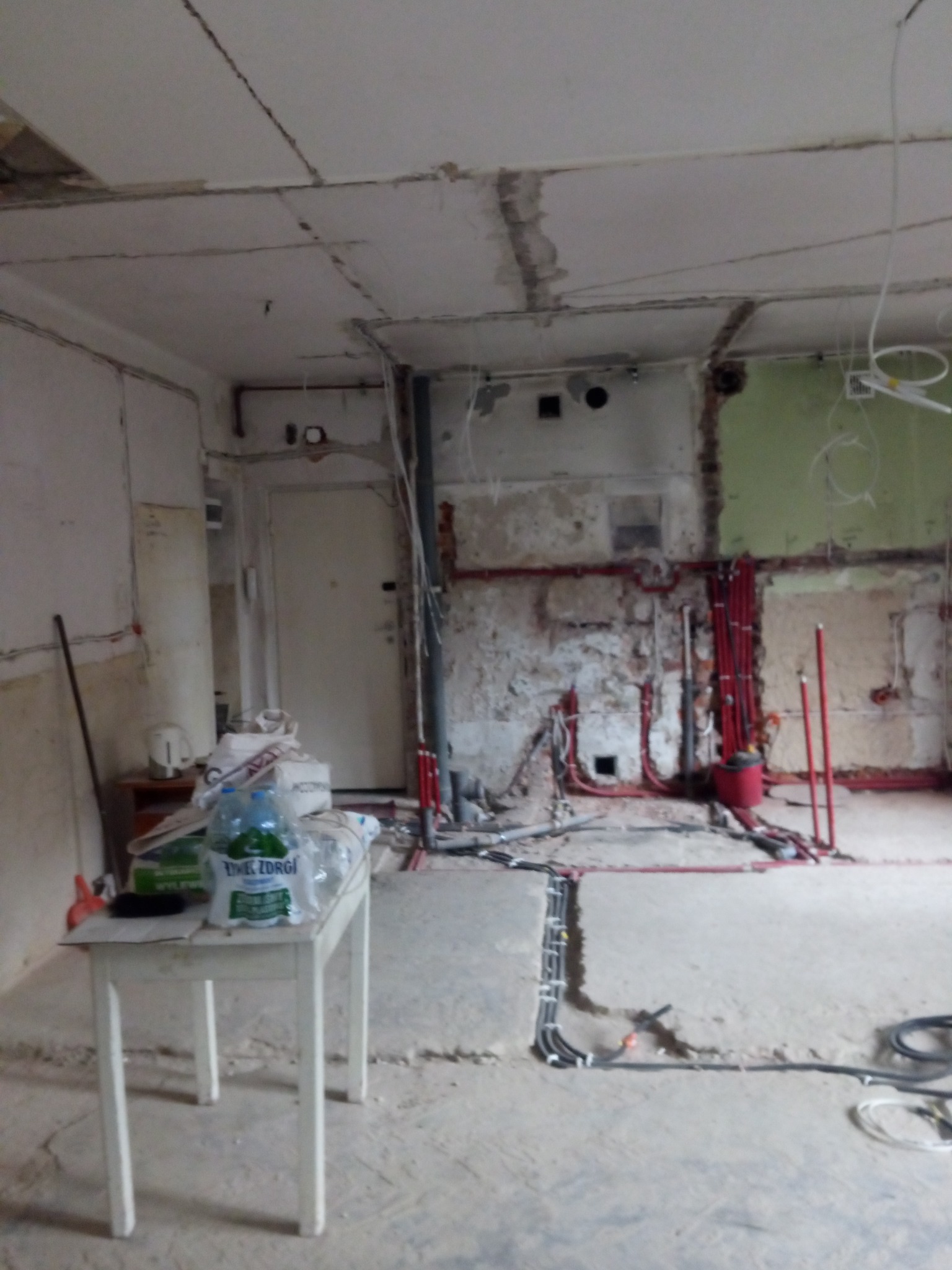 Wnętrze w trakcie remontu: odsłonięte instalacje elektryczne i hydrauliczne na ścianach, częściowo zerwana podłoga, prowizoryczny stolik z butelkami wody i narzędziami, surowy sufit z widocznymi...