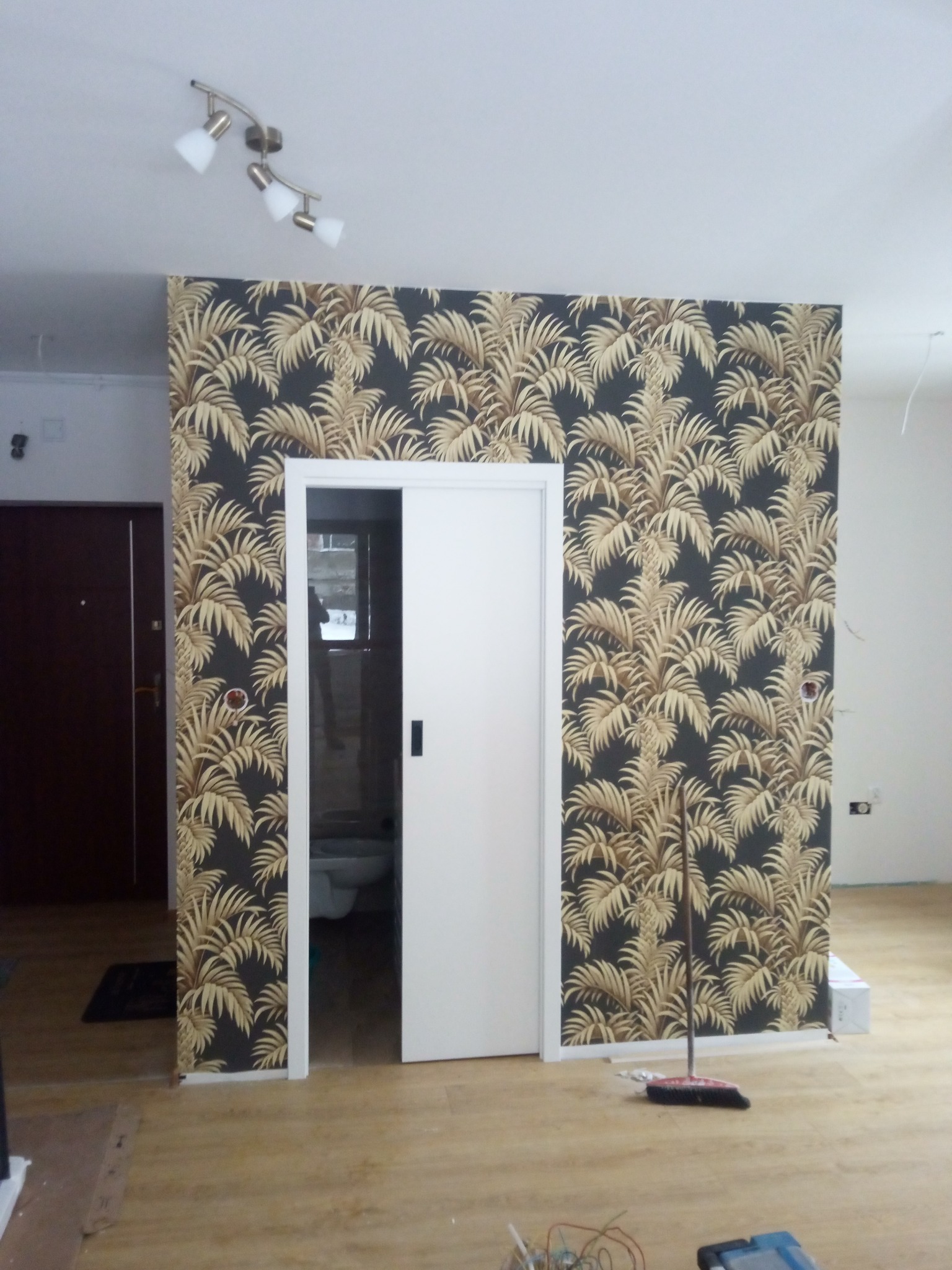Ściana z tapetą w motywy tropikalne z widocznymi drzwiami przesuwnymi i fragmentem łazienki w tle, pomieszczenie w trakcie prac wykończeniowych, widoczna miotła i narzędzia na podłodze.