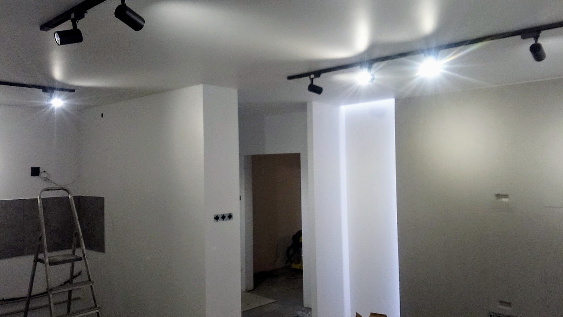 Wnętrze w trakcie remontu z białymi ścianami, widoczne szyny z reflektorami sufitowymi, drabina, przygotowane płyty oraz instalacja elektryczna w ścianie.