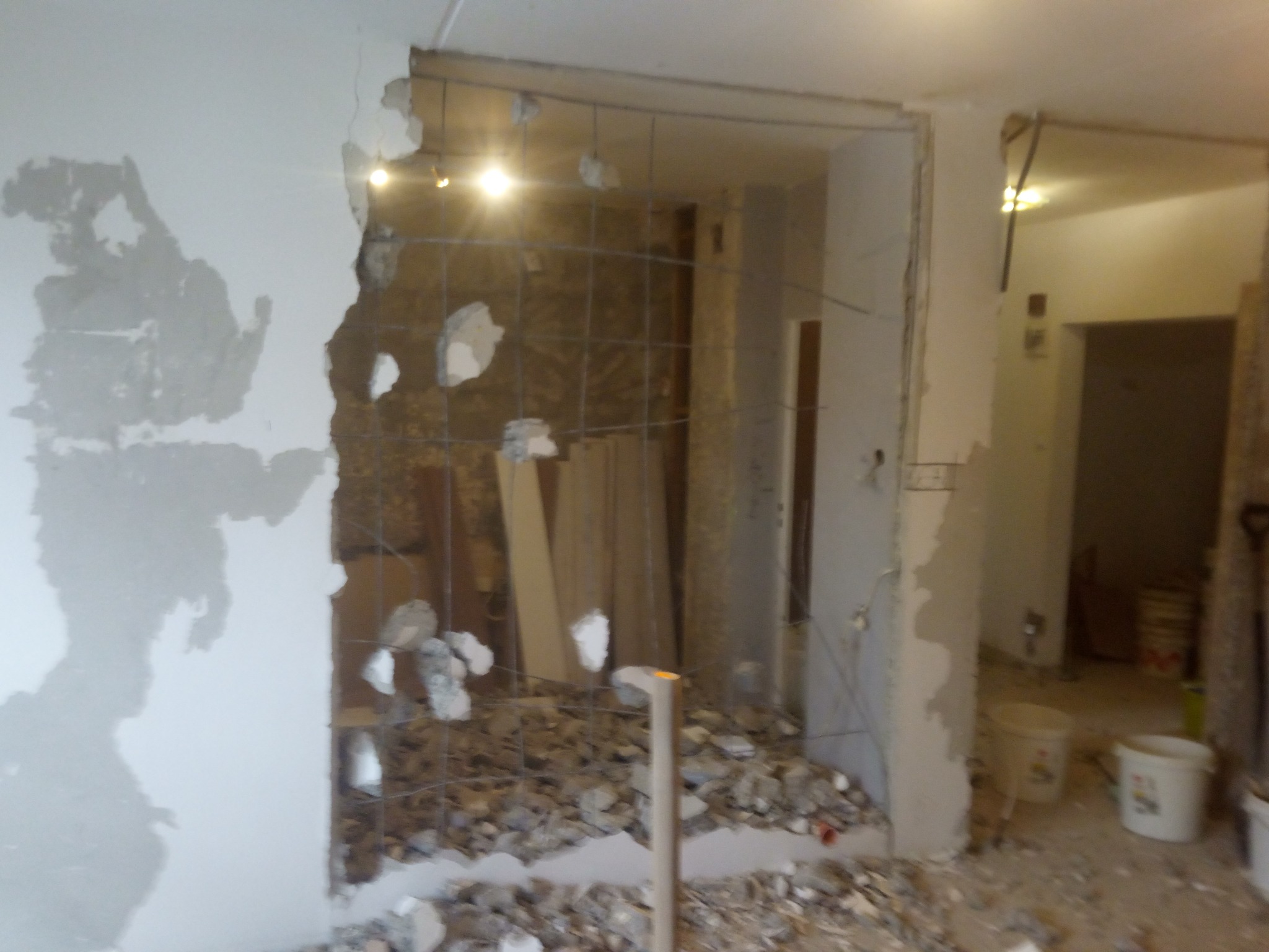 Demontaż ściany działowej z widocznymi fragmentami gruzu, odsłoniętą siatką zbrojeniową i resztkami starej zaprawy na ścianie, w tle widoczne puste pomieszczenie i wiaderka.