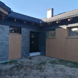 Elewacja domu jednorodzinnego z szarą okładziną kamienną i drewnianymi panelami, czarne drzwi wejściowe, widoczne okna z drewnianą stolarką, teren wokół domu w trakcie prac wykończeniowych.