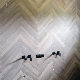 Ściana łazienki wyłożona płytkami w jodełkę, imitującymi drewno, z zamontowaną czarną armaturą i otworem na gniazdko.