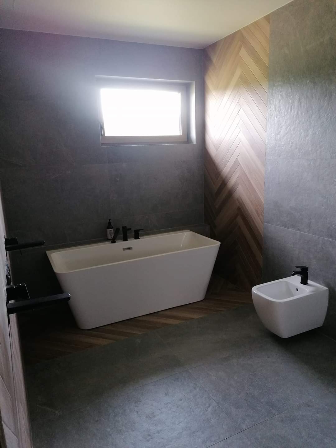 Nowoczesna łazienka z wolnostojącą białą wanną, bidetem i szarą płytką imitującą kamień, akcentowana ścianą z drewnianą jodełką.