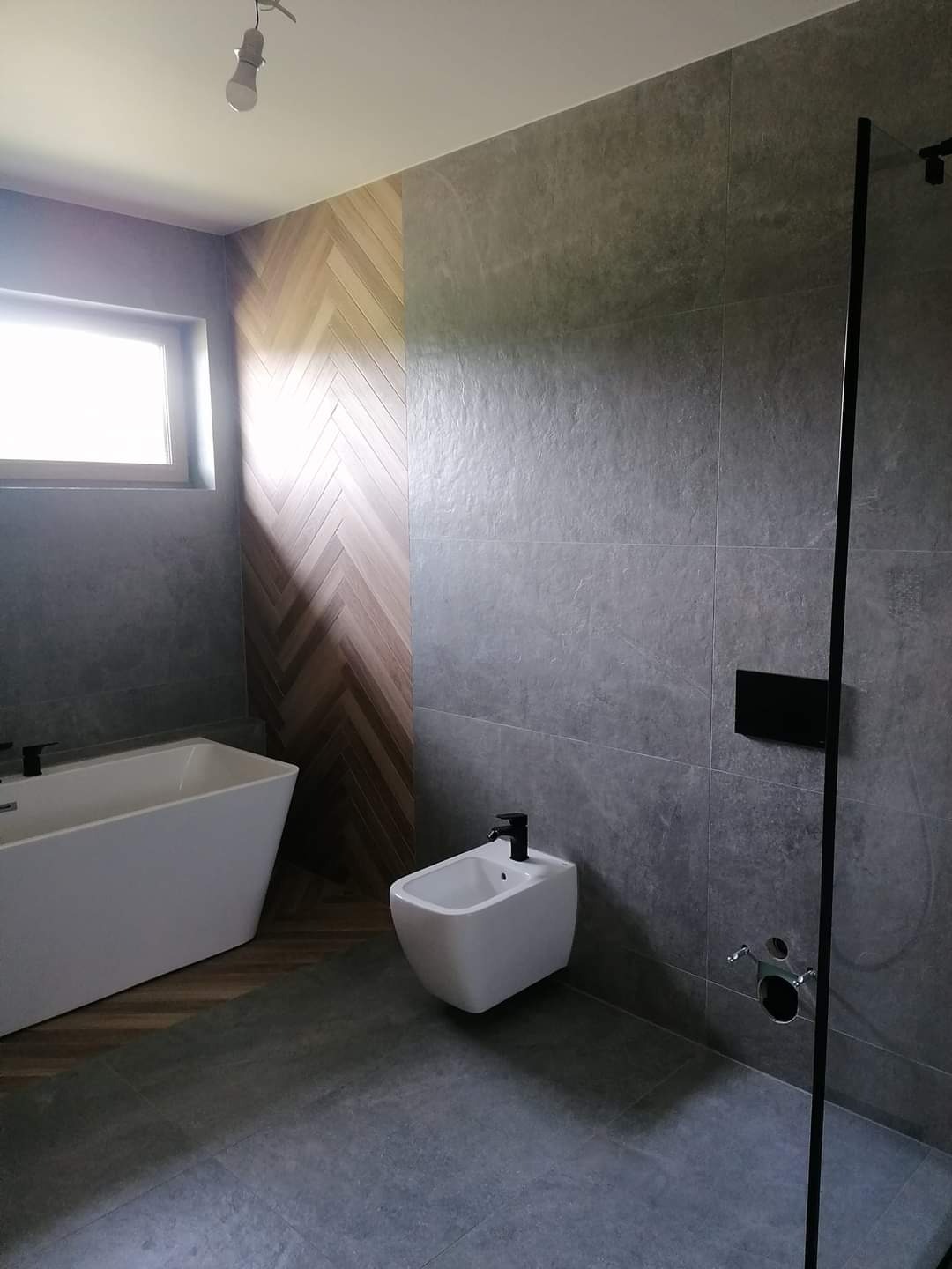 Nowoczesna łazienka z szarymi płytkami, drewnianą ścianą ułożoną w jodełkę, białą wanną i bidetem oraz czarnymi akcentami, takimi jak bateria i rama kabiny prysznicowej.
