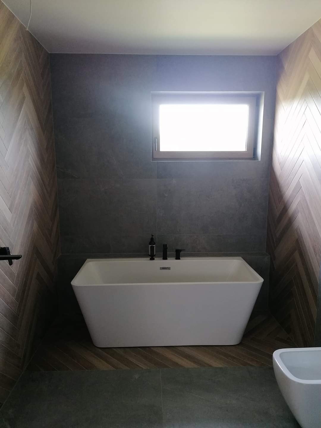Nowoczesna łazienka z białą, prostokątną wanną, szarą płytką ścienną, drewnopodobną okładziną w jodełkę na ścianach bocznych i małym oknem wnękowym w górnej części ściany.