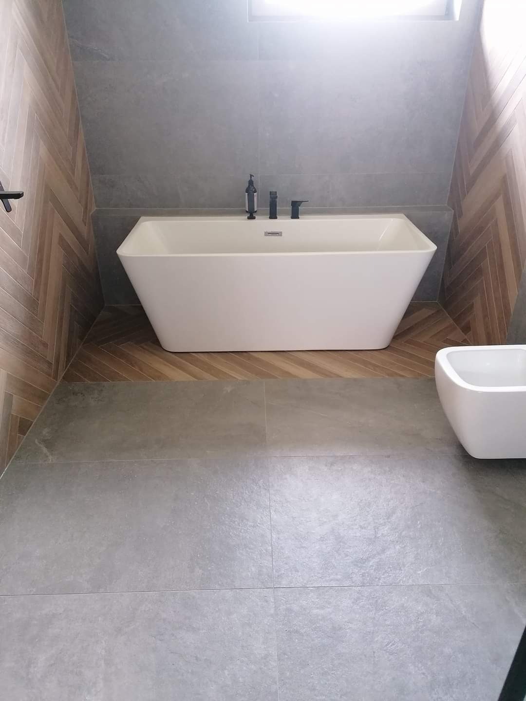 Nowoczesna łazienka z wolnostojącą białą wanną, czarną armaturą i szarą płytką podłogową oraz drewnianą mozaiką na ścianach.