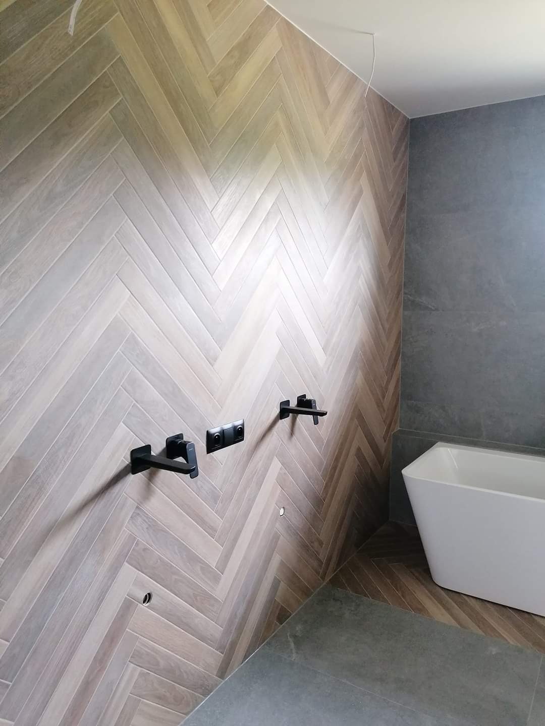 Ściana łazienki wyłożona płytkami imitującymi drewno ułożonymi we wzór jodełki, z zamontowaną czarną armaturą i widoczną białą wanną.