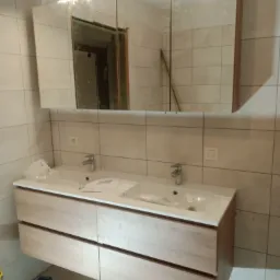 Nowoczesna szafka łazienkowa z dwiema umywalkami i lustrem z oświetleniem, widoczne elementy instalacji hydraulicznej, płytki ceramiczne na ścianach.