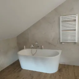 Biała wolnostojąca wanna w łazience na poddaszu z panelami imitującymi drewno, grzejnik drabinkowy na ścianie z marmurkowym wzorem.