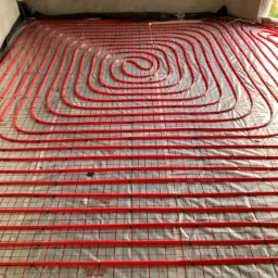 Czerwone rury ogrzewania podłogowego ułożone spiralnie na folii z siatką, widok z góry na surowy stan posadzki w trakcie instalacji.