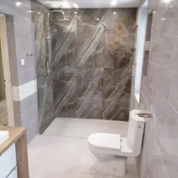 Nowoczesna łazienka z kabiną prysznicową typu walk-in, wykończona płytkami imitującymi marmur w odcieniach szarości i brązu, biała ceramika sanitarna: toaleta i bidet, umywalka z drewnianą szafką.
