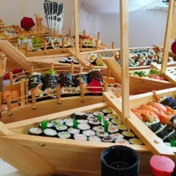 Dekoracyjny stół z sushi w drewnianych łodziach, różnorodne rolki i nigiri, stosy pałeczek do sushi z nadrukiem, czerwone róże jako ozdoby.